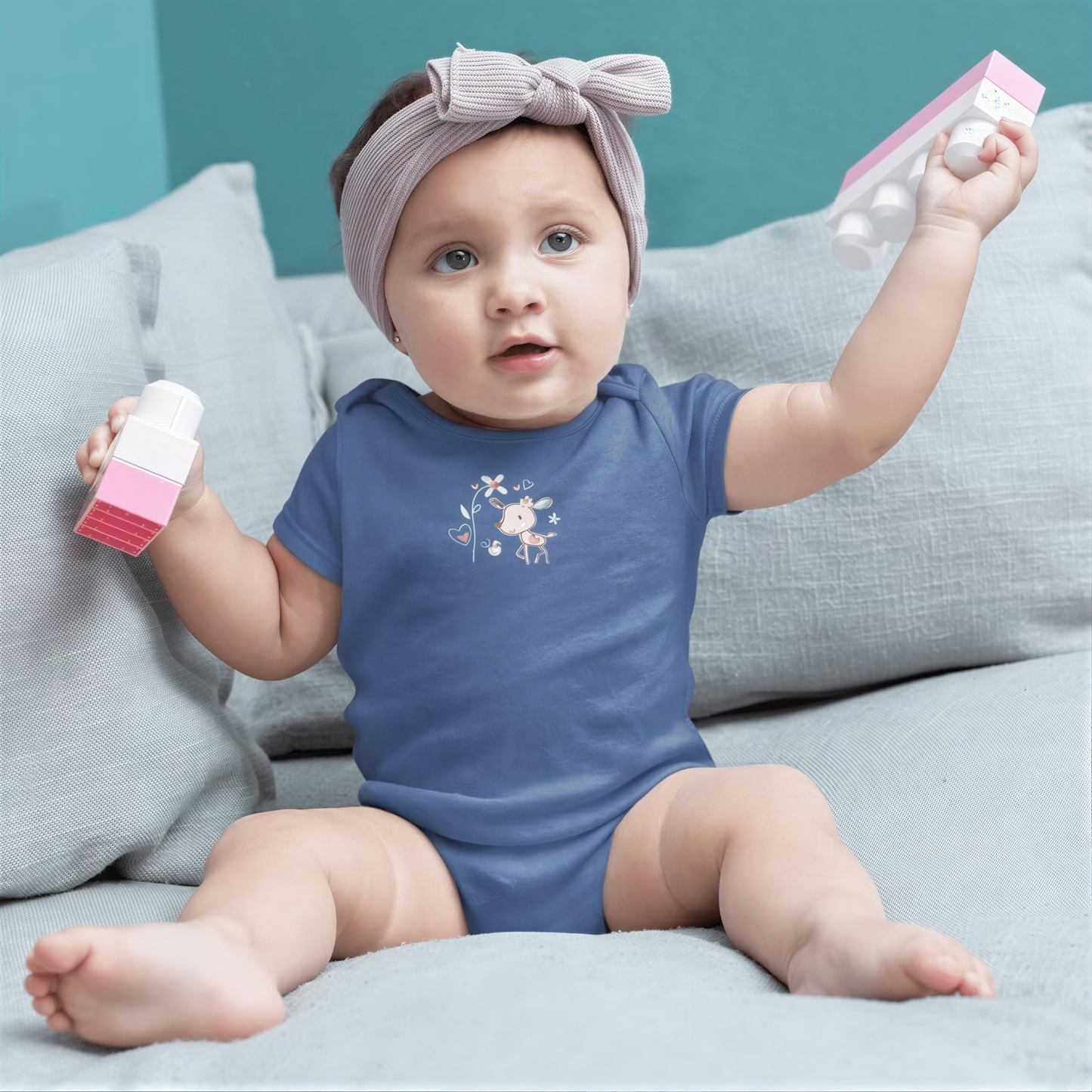 A baby girl wearing a blue onesie and a gray headband is sitting on a gray couch, holding a pink and white toy.