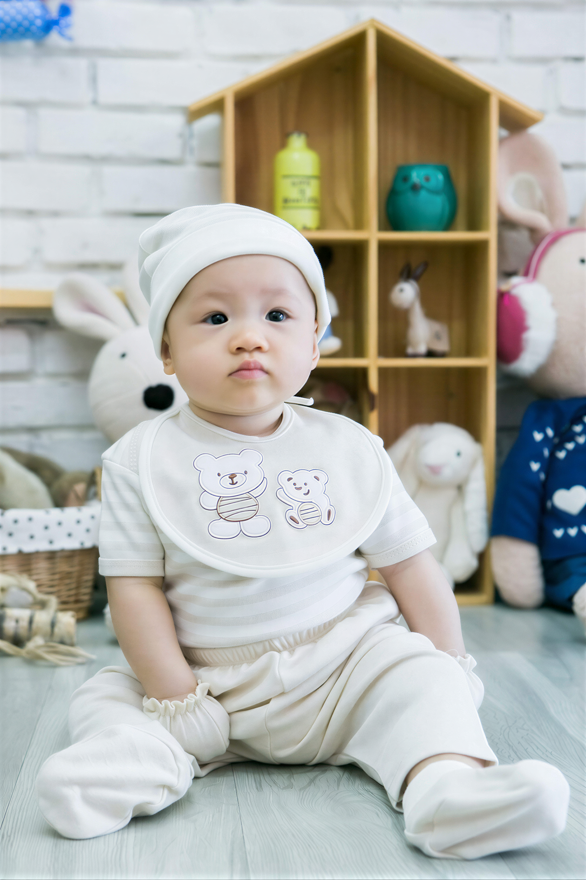 A baby boy wearing a white outfit and bib sits on the floor in front of a wooden toy house, surrounded by various stuffed animals.