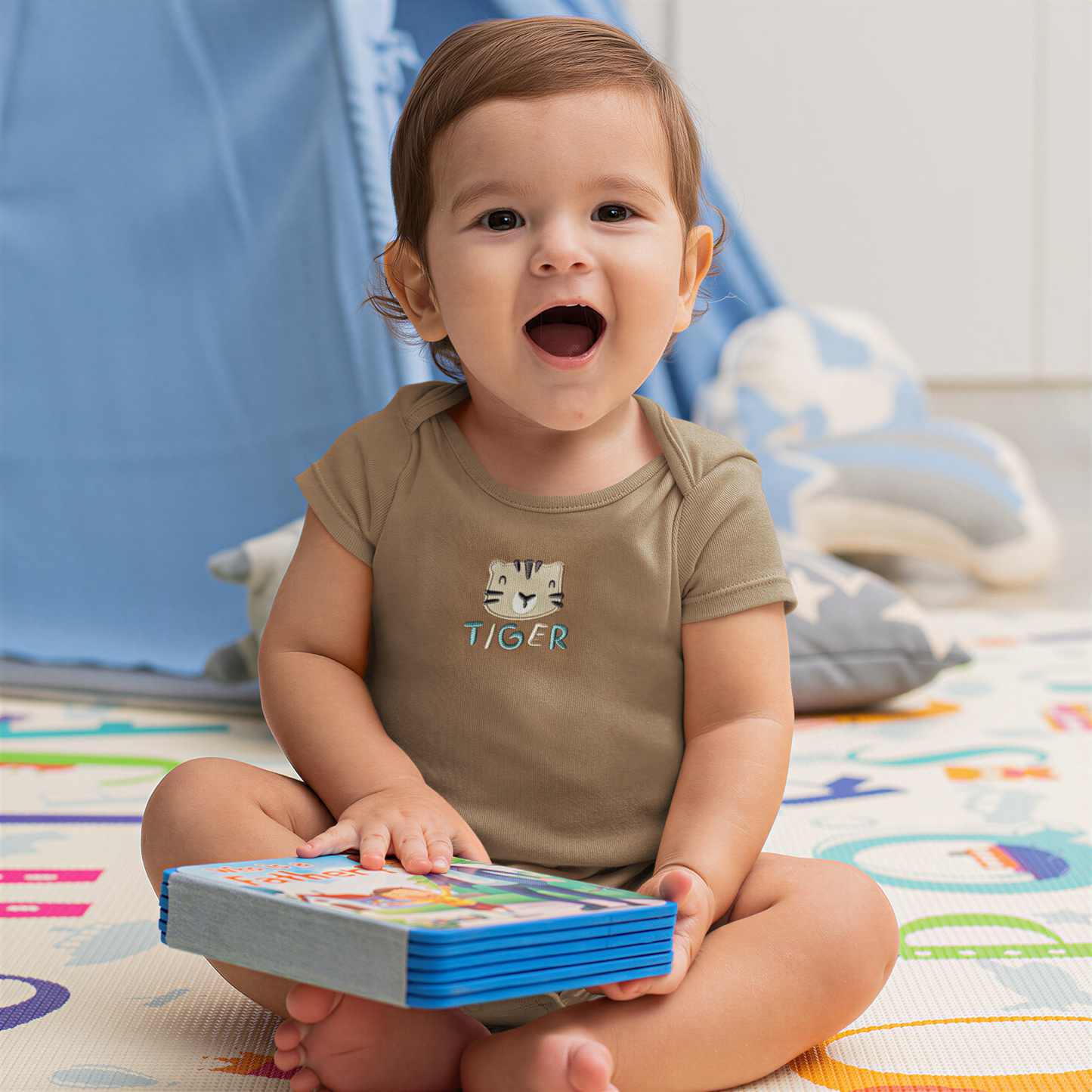 A smiling baby boy sitting on the floor, holding a book and looking at the camera.