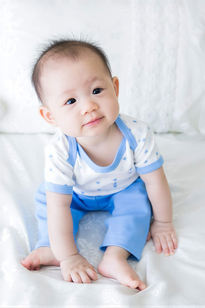 A smiling baby boy wearing a white and blue outfit sits on a white bedspread.