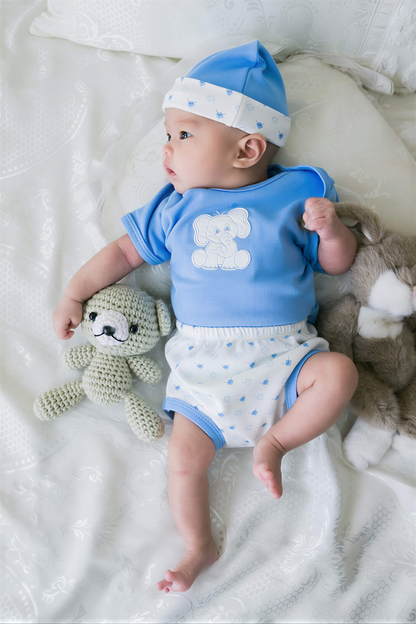 A baby wearing a blue outfit with a white polka dot pattern lies on a white bed, holding a small crocheted teddy bear.